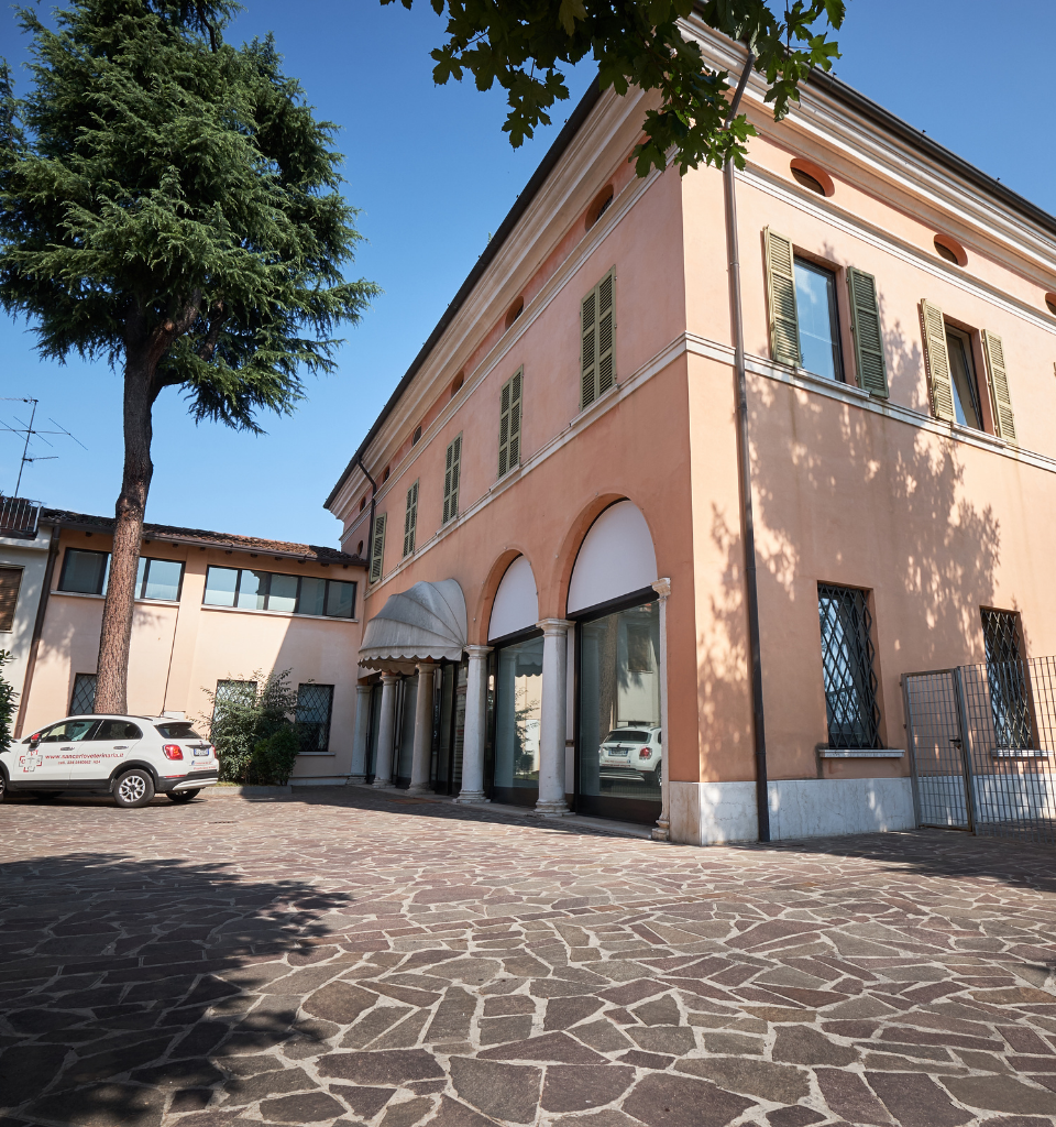 Un grande edificio color pesca con portali ad arco e finestre verdi chiuse si trova accanto a un albero e a due auto bianche parcheggiate in un cortile lastricato di pietra, sotto un cielo azzurro e limpido.